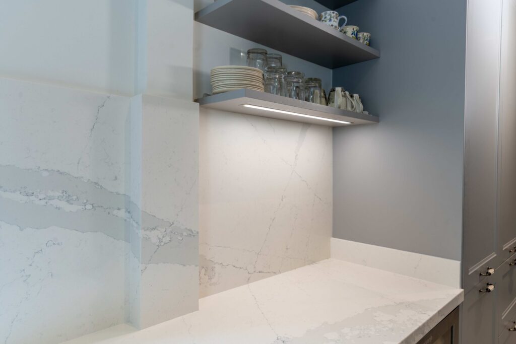 A closeup of the quartz worktop and niche cladding blending together underneath a section of open shelving in a cottage kitchen.
